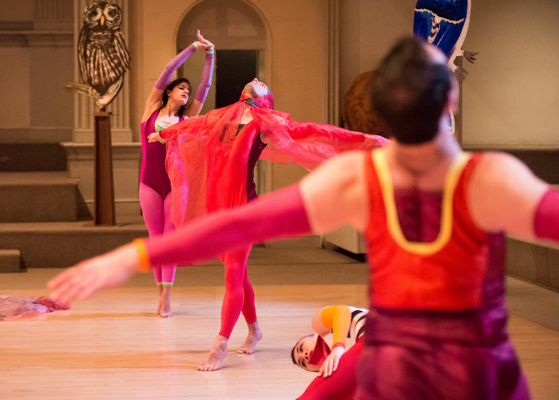 Dancers in purple and red costumes. A dancer in the center wears a gauzy scarf over her head and that drapes over her arms like bird's wings.